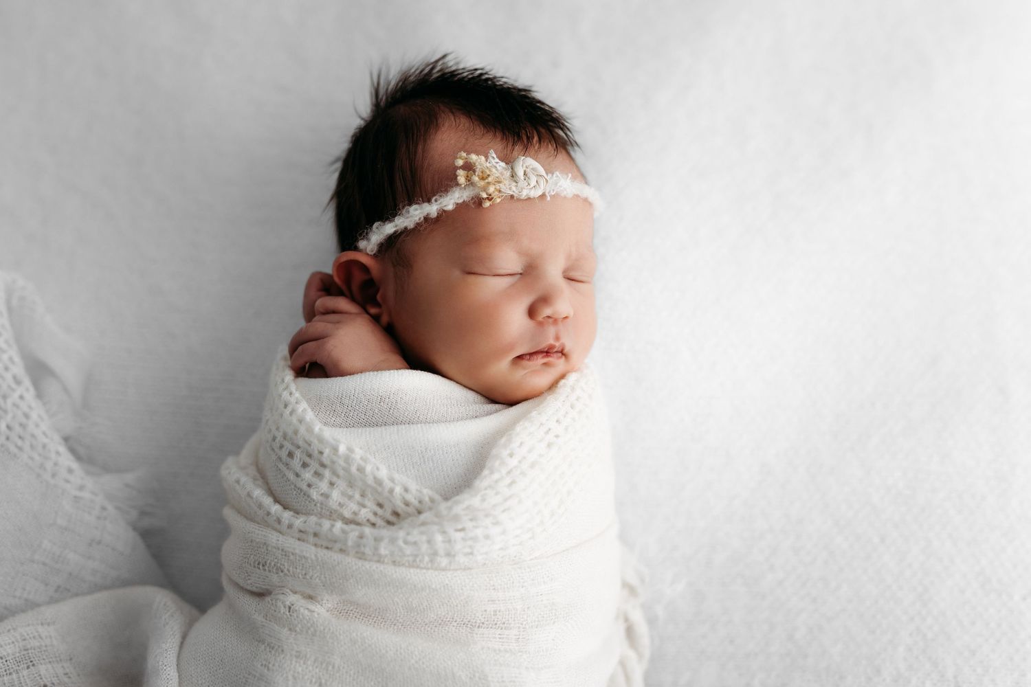Newborn baby wrapped in soft white blanket wearing delicate pearl headband during professional studio photoshoot. Rashinphotography-Nova DMV