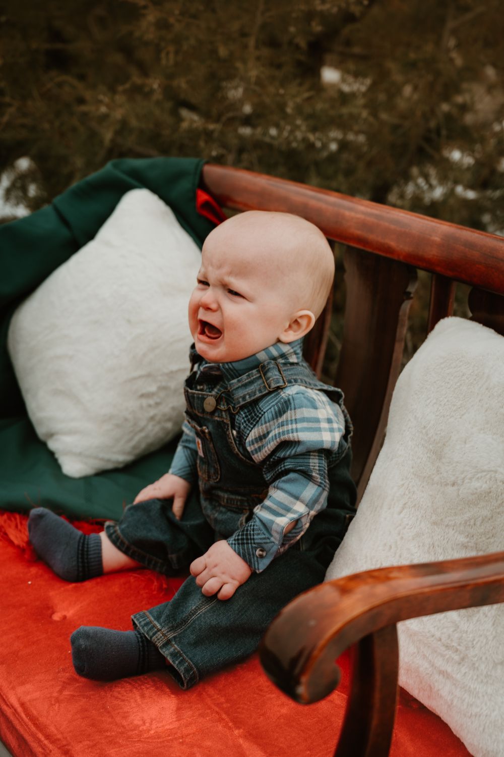 Baby in plaid shirt and overalls laughing while sitting on red cushions outdoors.
