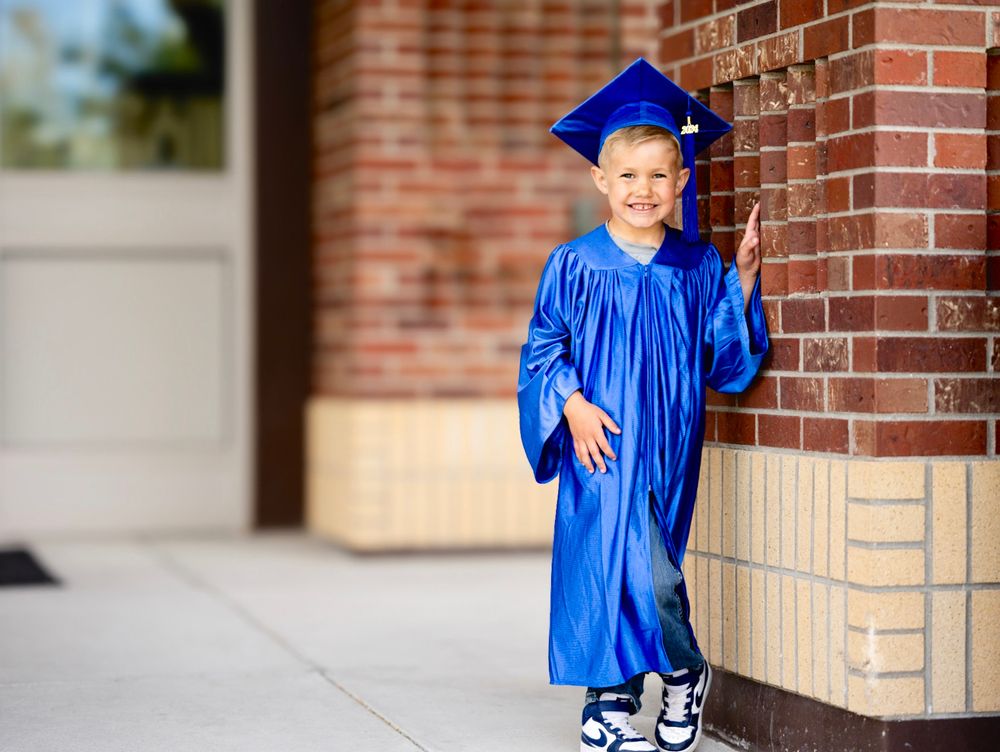 Young graduate in bright blue cap and gown poses by brick school building.