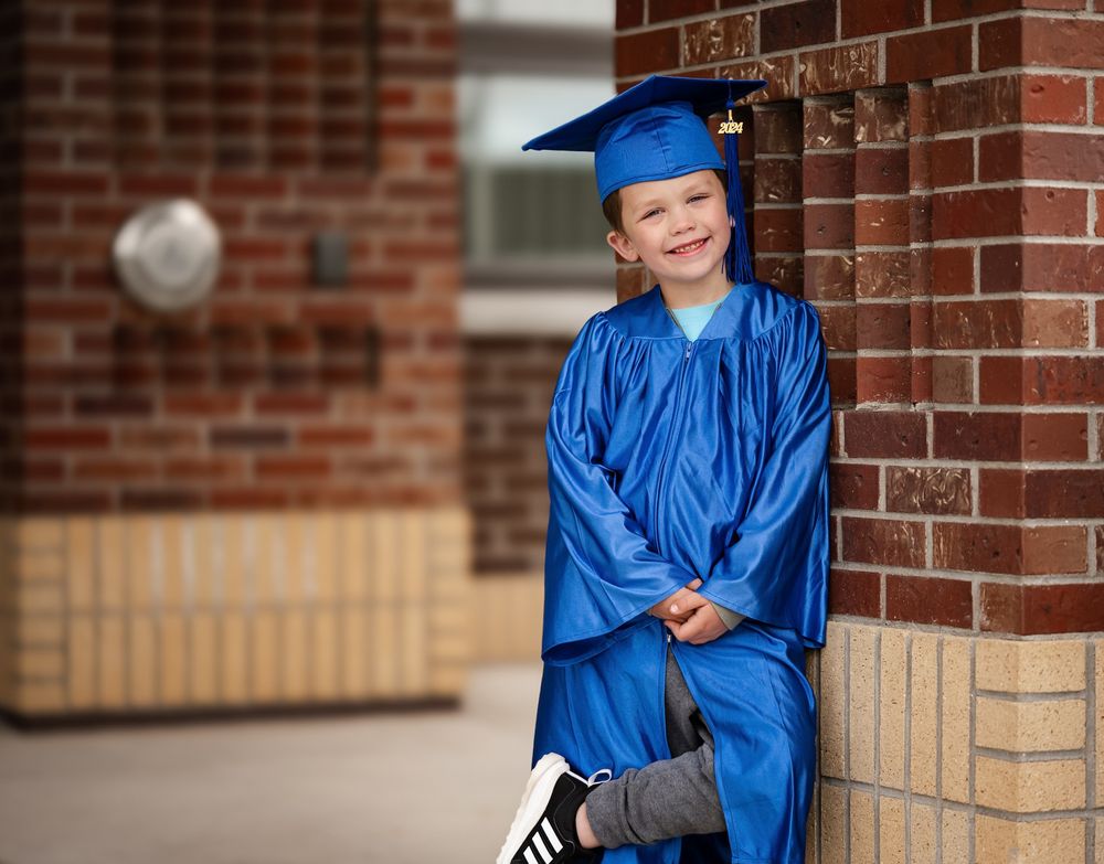 Graduate in royal blue cap and gown poses against brick wall.