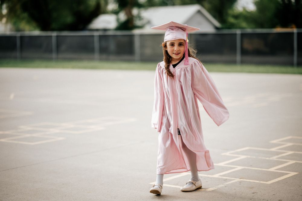 Person in pink graduation gown plays hopscotch on playground.