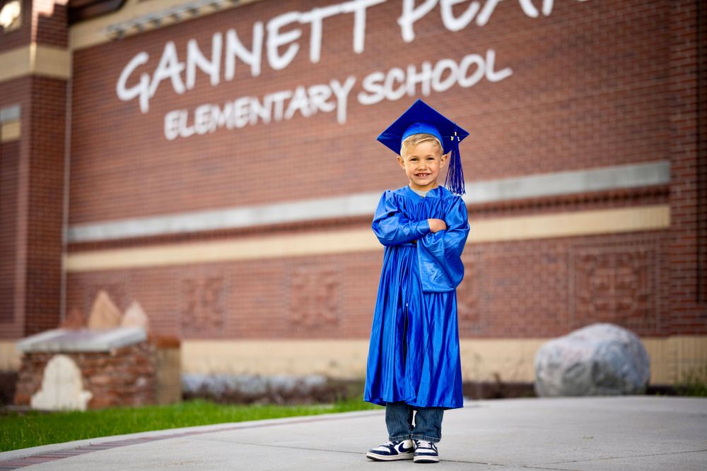 A graduate in blue cap and gown poses in front of a brick elementary school building.