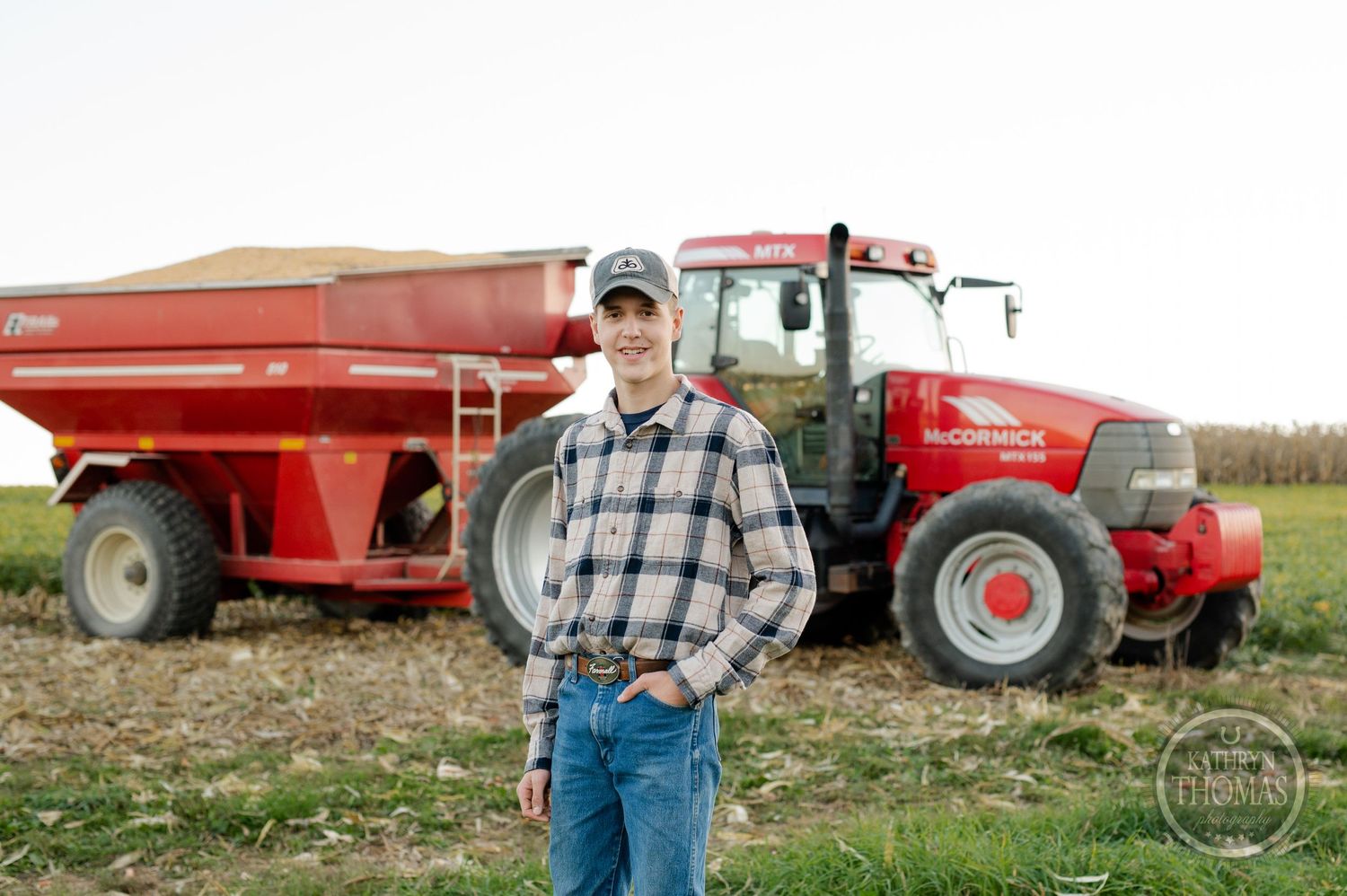 A farmer poses in front of a red tractor and grain cart in an agricultural field.