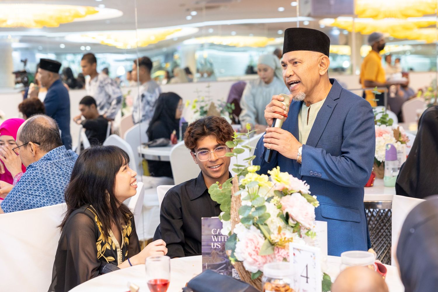 A speaker addresses wedding guests during a reception in a brightly lit venue.