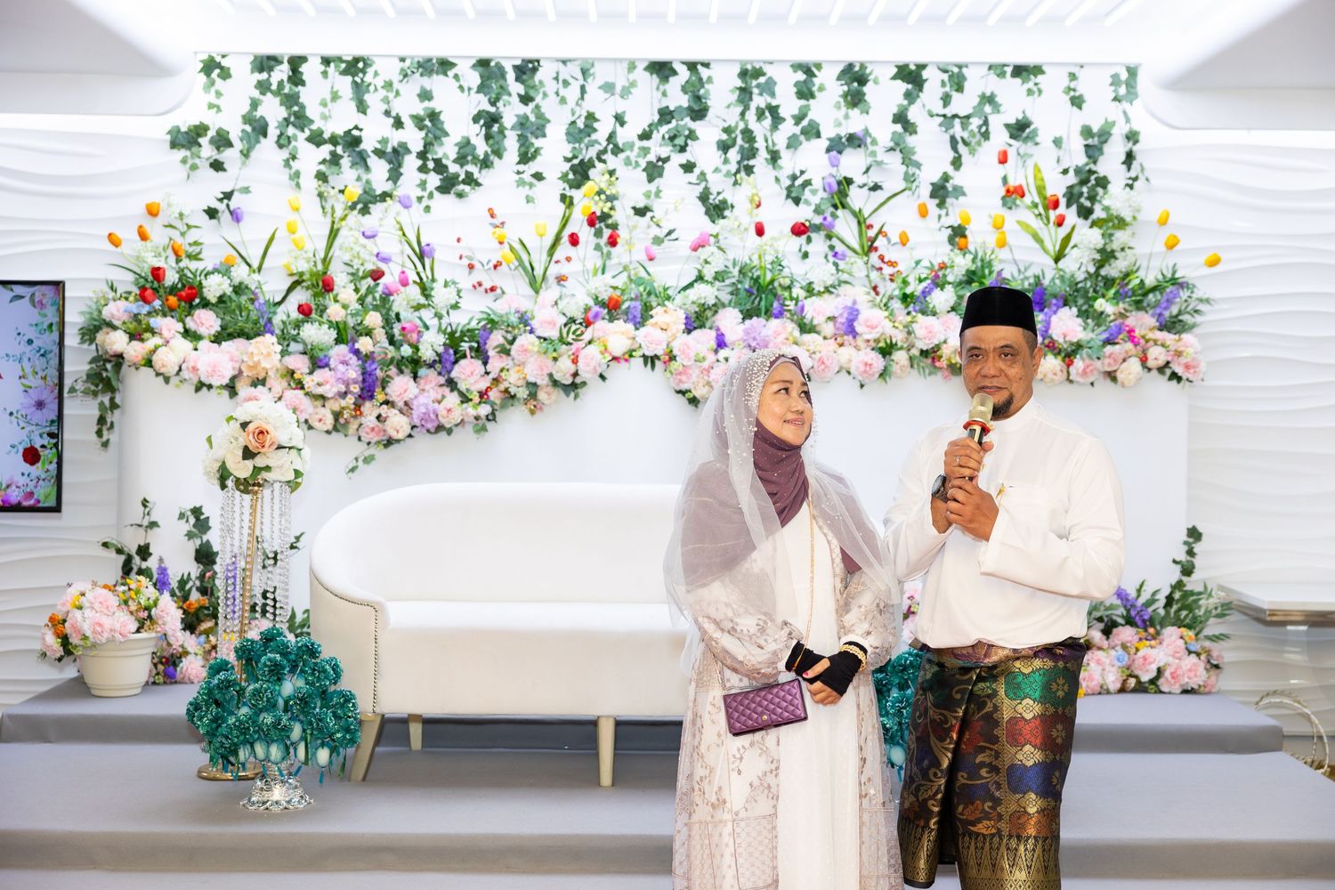 Couple in traditional Malay wedding attire standing against floral backdrop.