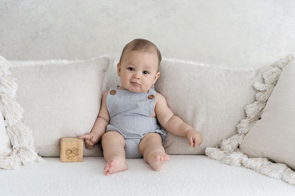 Baby Photography Melbourne, a baby wearing a gray romper sits on a white couch with decorative throw pillows and a wooden block toy.