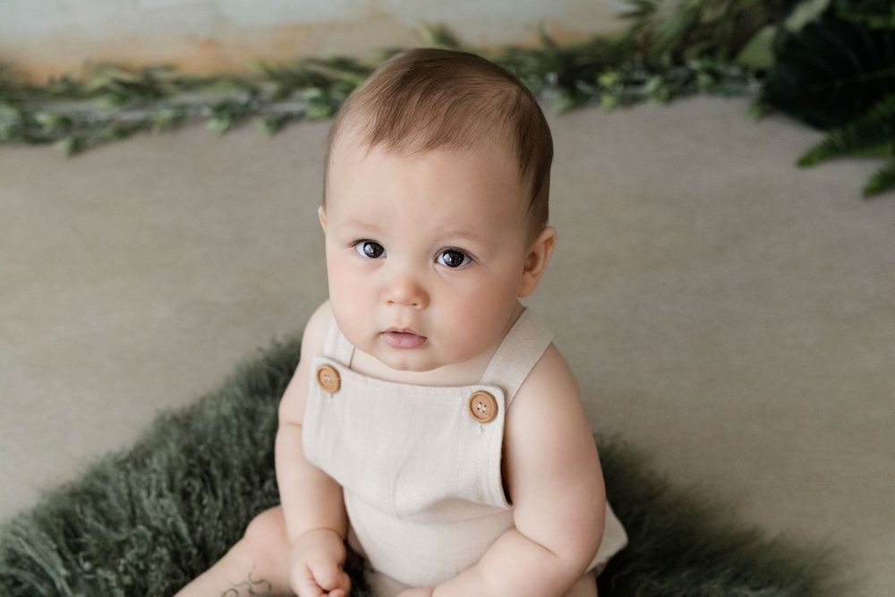 Baby in white onesie sitting on floor with greenery background.