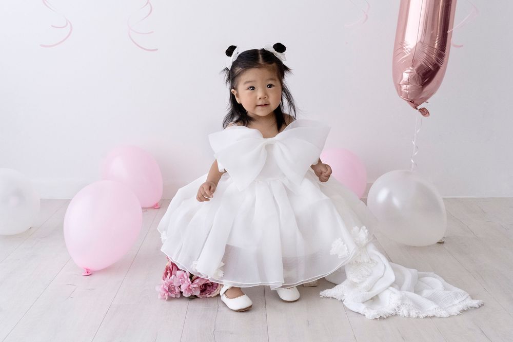 A child in a white dress poses against a pink and white balloon backdrop for a birthday photoshoot.