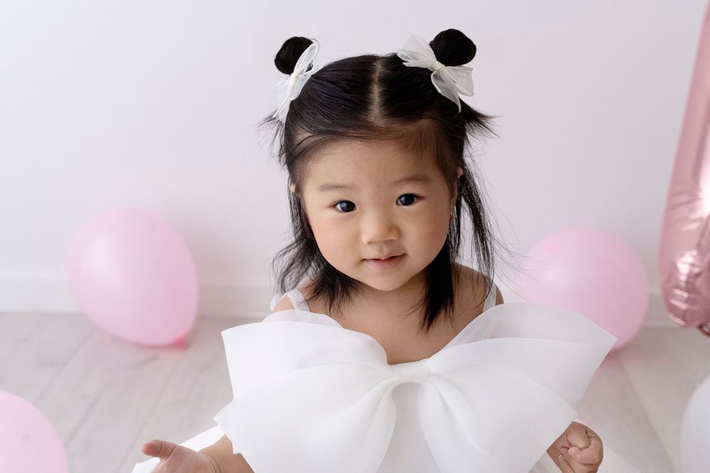 A child wearing a white dress smiles while surrounded by pink and white balloons.