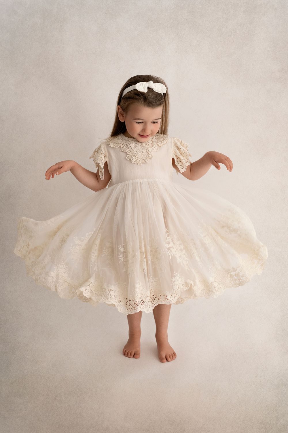 Girl twirling in delicate ivory lace dress against light background.