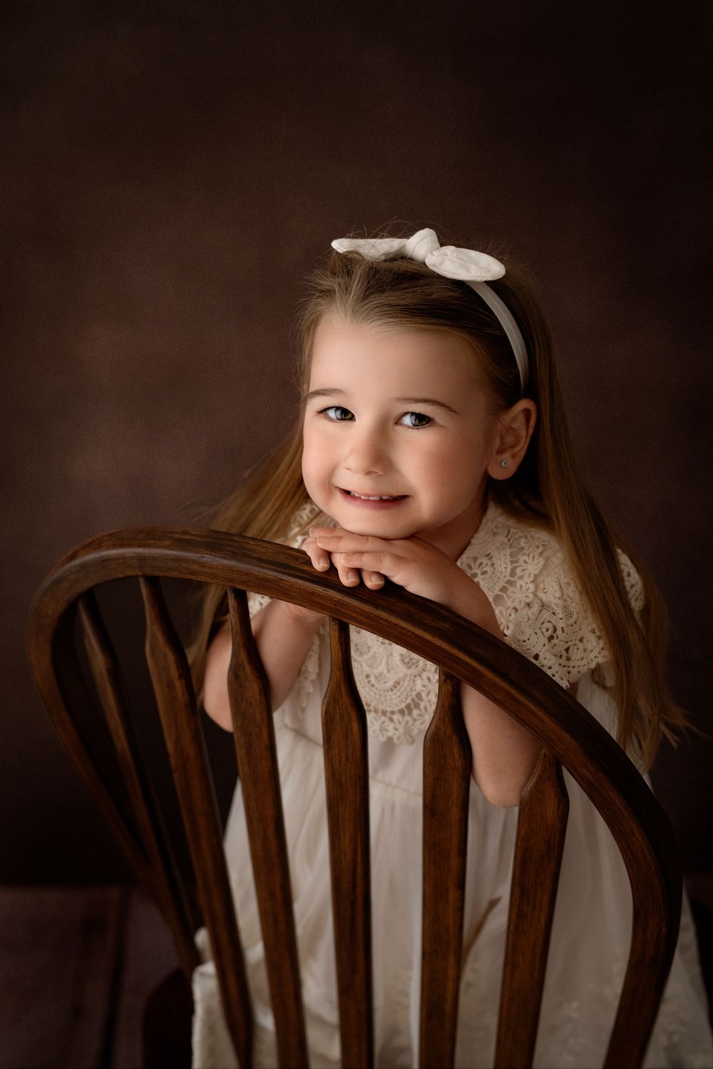 Child peeking playfully over wooden chair back in white lace dress.