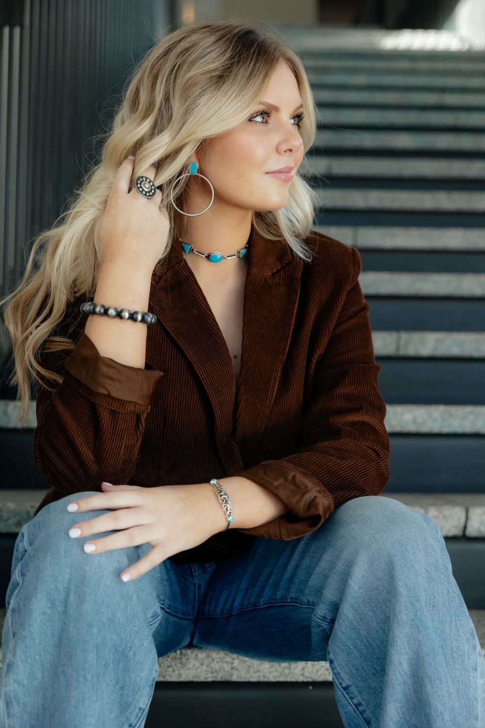Blonde in brown corduroy blazer and turquoise jewelry sits thoughtfully on concrete steps wearing blue jeans.