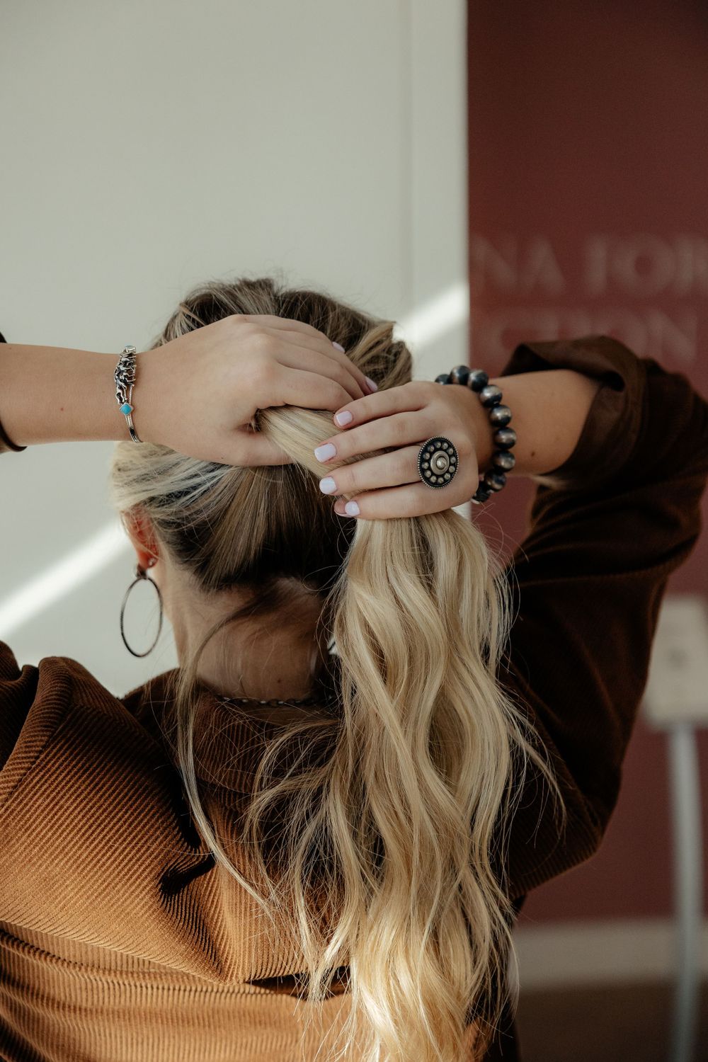 Person with long blonde hair wearing a brown sweater styles their hair while wearing boho-style jewelry and rings.