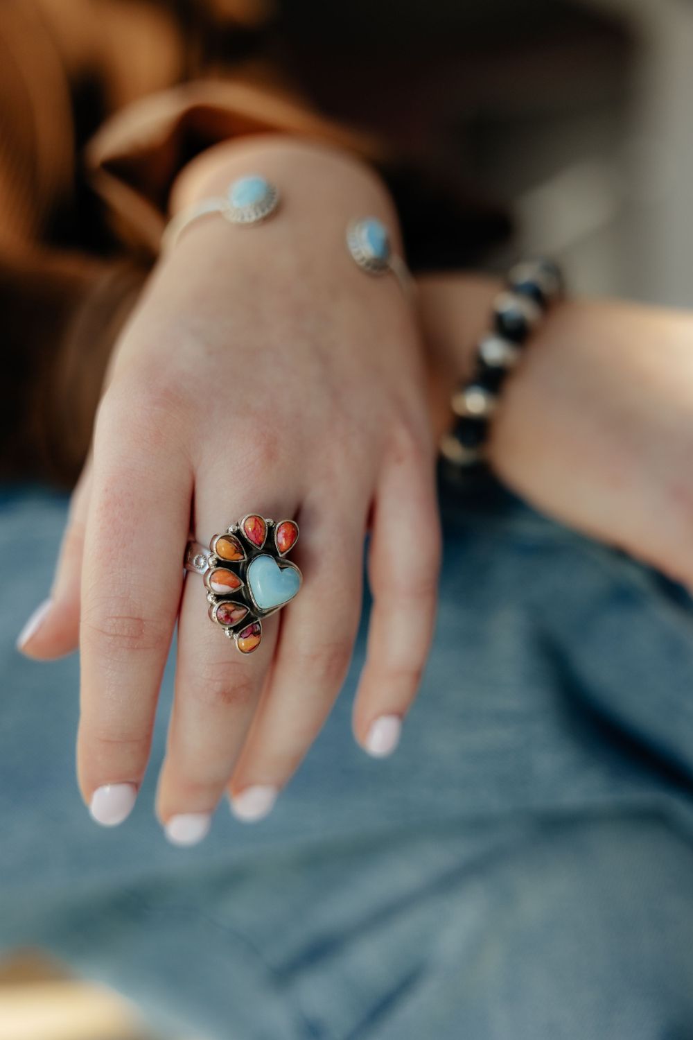 A hand wearing a bohemian turquoise and coral statement ring with white nail polish against denim fabric.