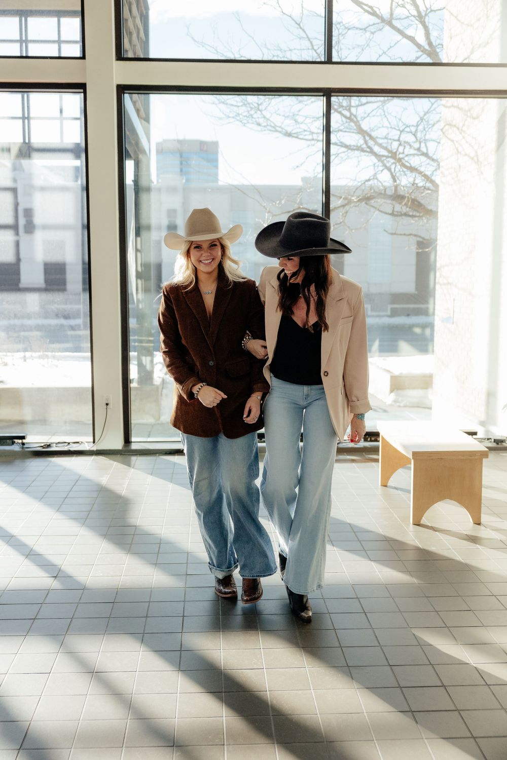 Two people in western style outfits with cowboy hats, blazers and jeans walking together in a sunlit indoor space.
