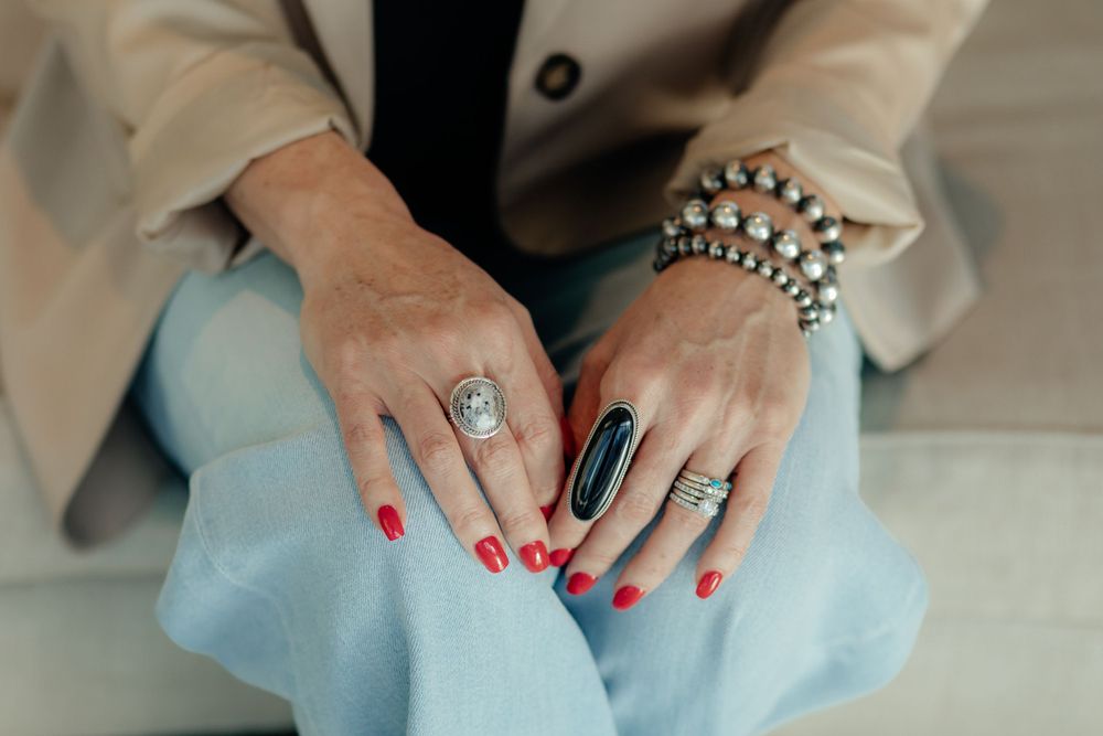 Close-up of hands wearing multiple rings and bracelets resting on light blue fabric with red nail polish.