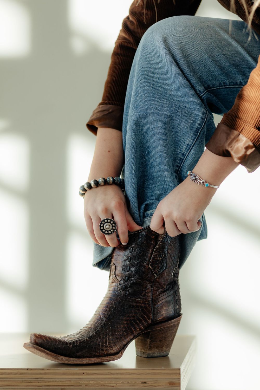 Person wearing brown western boots, jeans, and bracelets poses with foot elevated on wooden surface in artistic lighting.