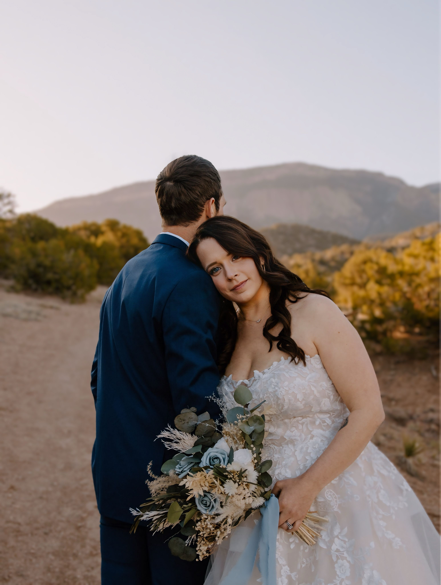 Newlyweds share an intimate moment against a mountainous backdrop with a rustic bouquet at sunset.
