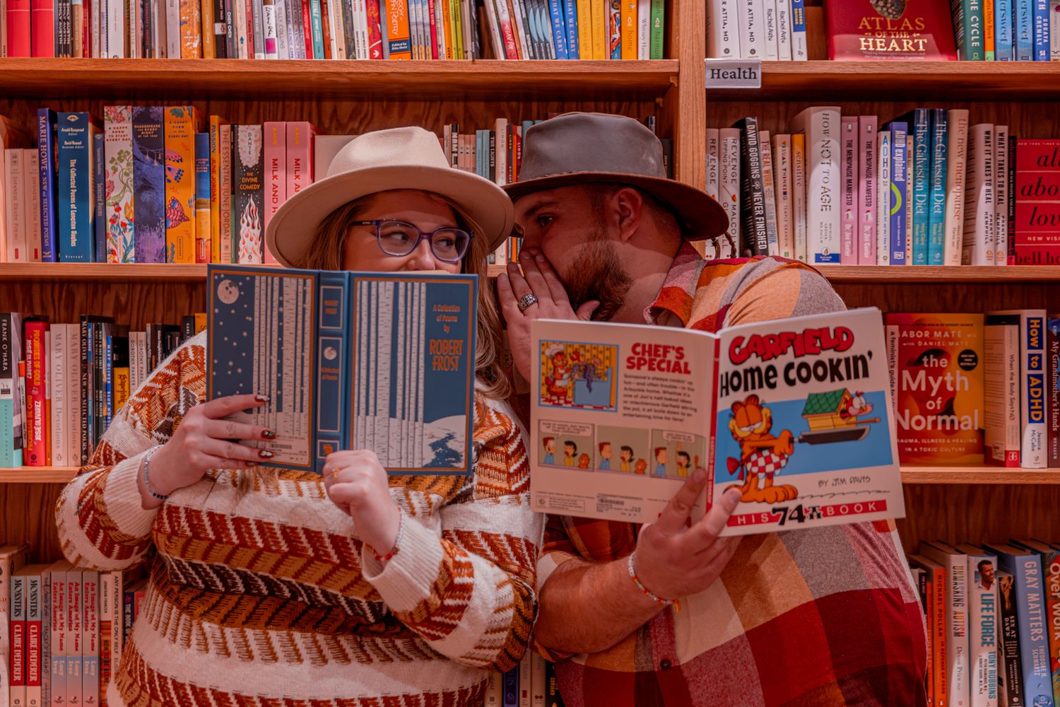 Two people wearing hats reading children's books in front of a crowded bookshelf in a library or bookstore.