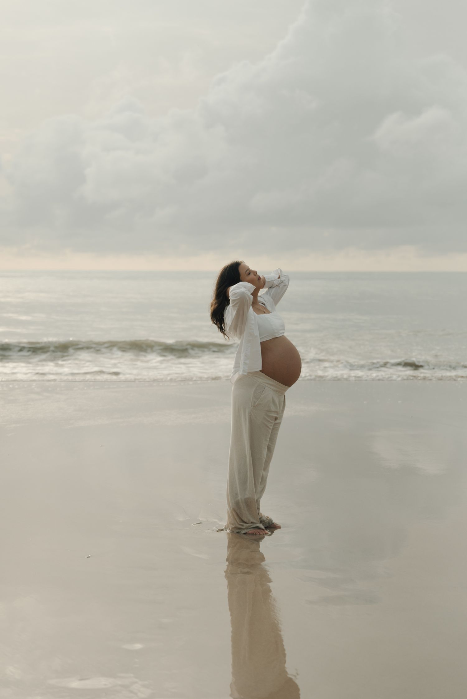 Expectant mother in white clothing stands reflectively on a beach at sunset.