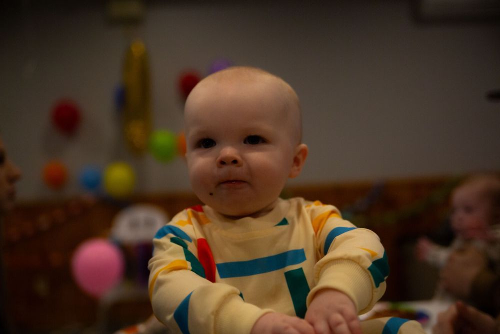A small baby wearing a yellow outfit with teal overalls sits surrounded by colorful play toys and balloons.