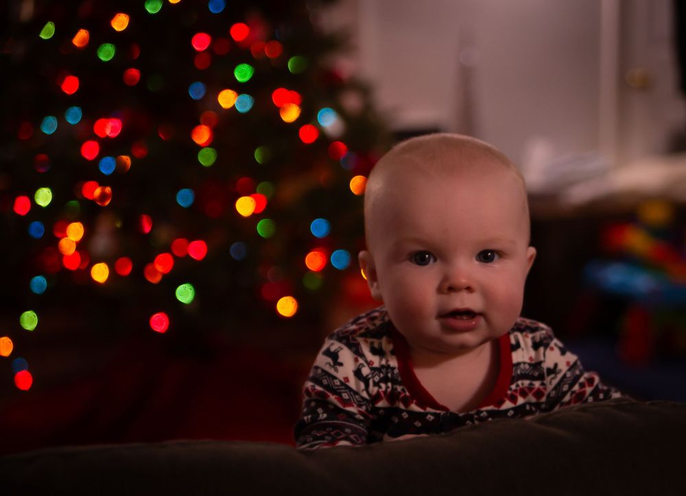 Baby in patterned sweater looking ahead with festive Christmas lights behind.