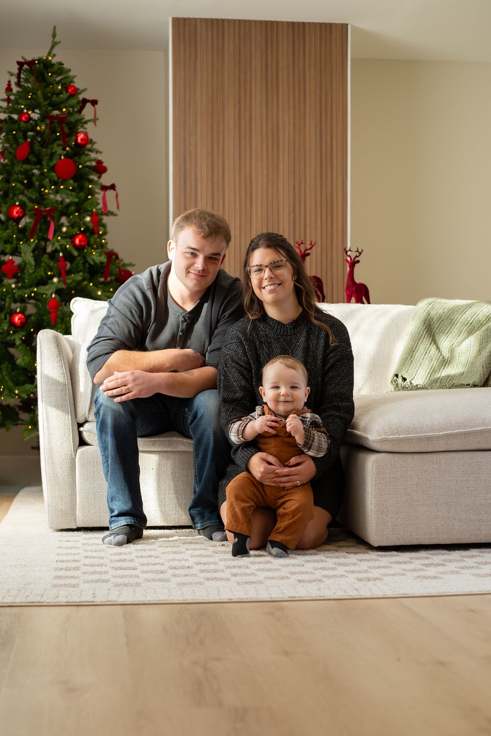 A warm family holiday photo on a white couch with a beautifully decorated Christmas tree and wooden accent wall.