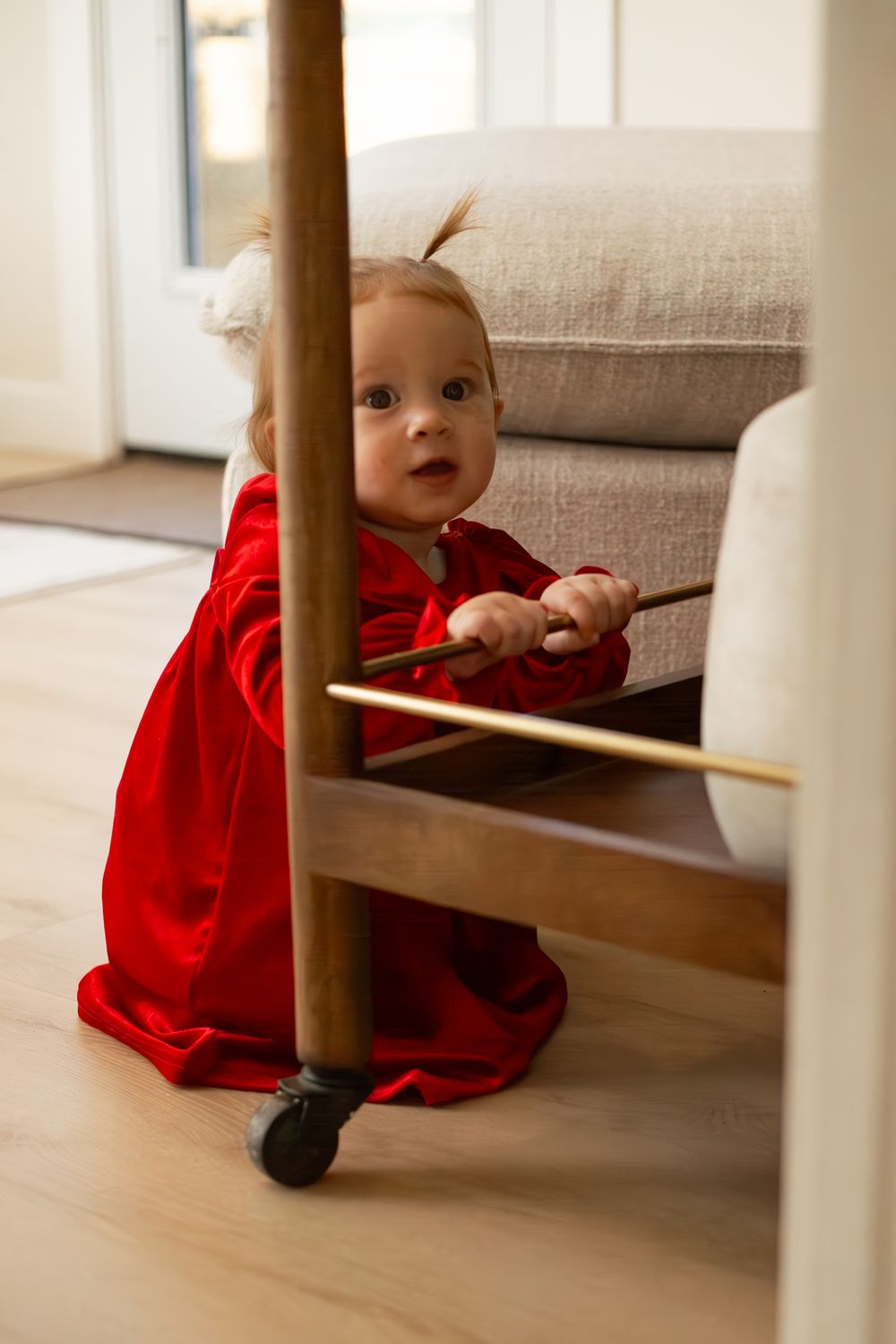 A small child in a bright red velvet dress peeks around a wooden furniture piece in a cozy living room.