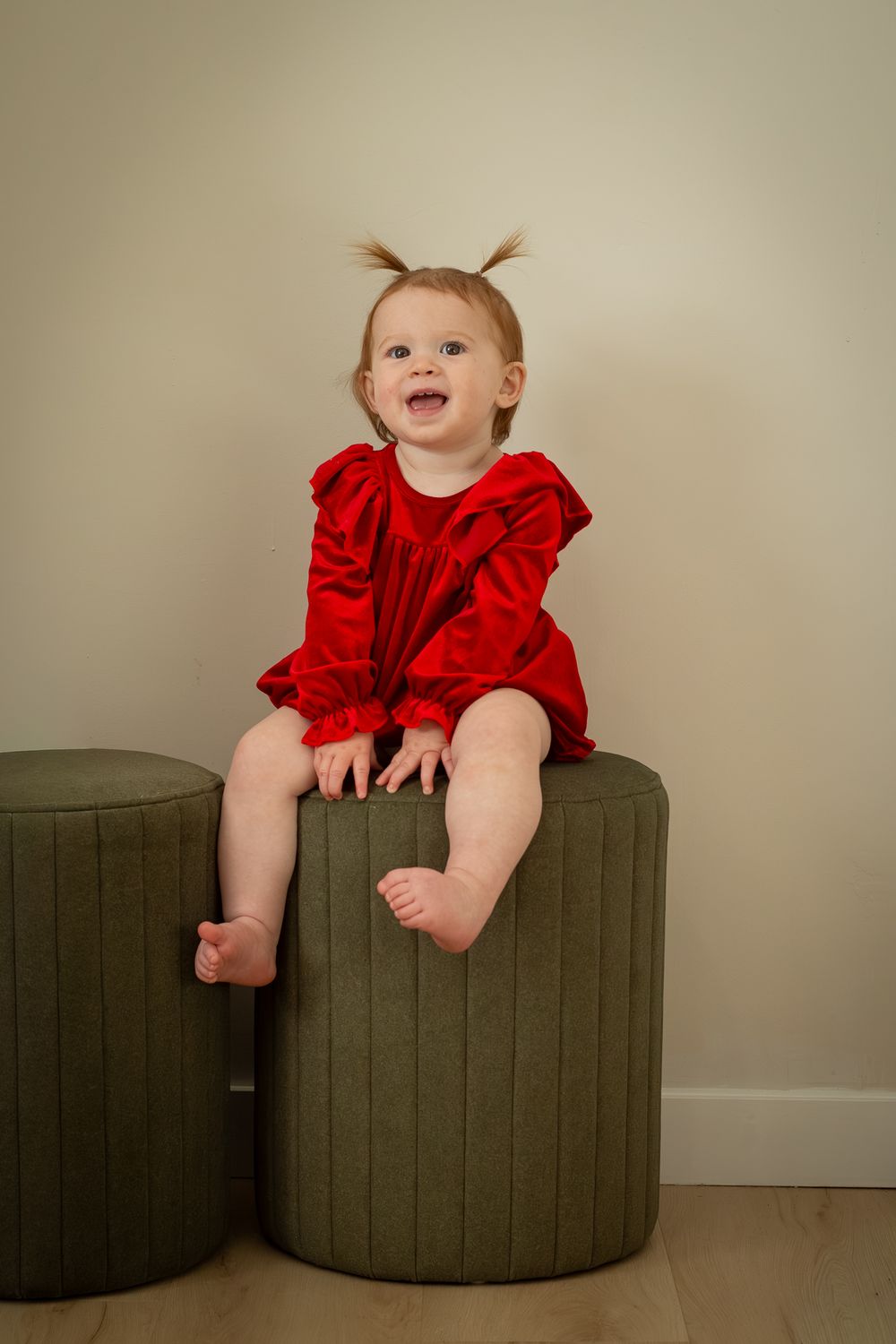 A small child in a festive red dress sits atop a dark green ottoman, displaying joyful expression and pigtails.