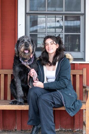 A happy black shaggy dog and person share a joyful moment on a wooden bench by a red building.