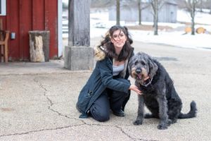A large black furry dog sits next to a red building during winter.