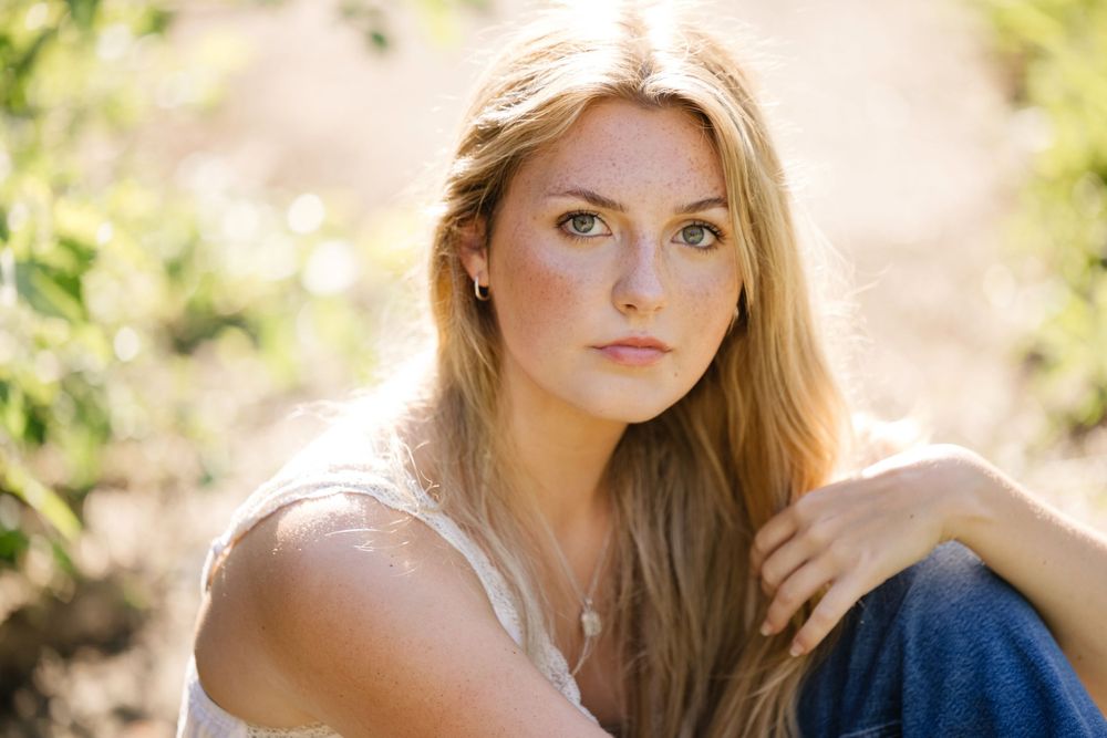 Natural light portrait against green foliage wearing white top.