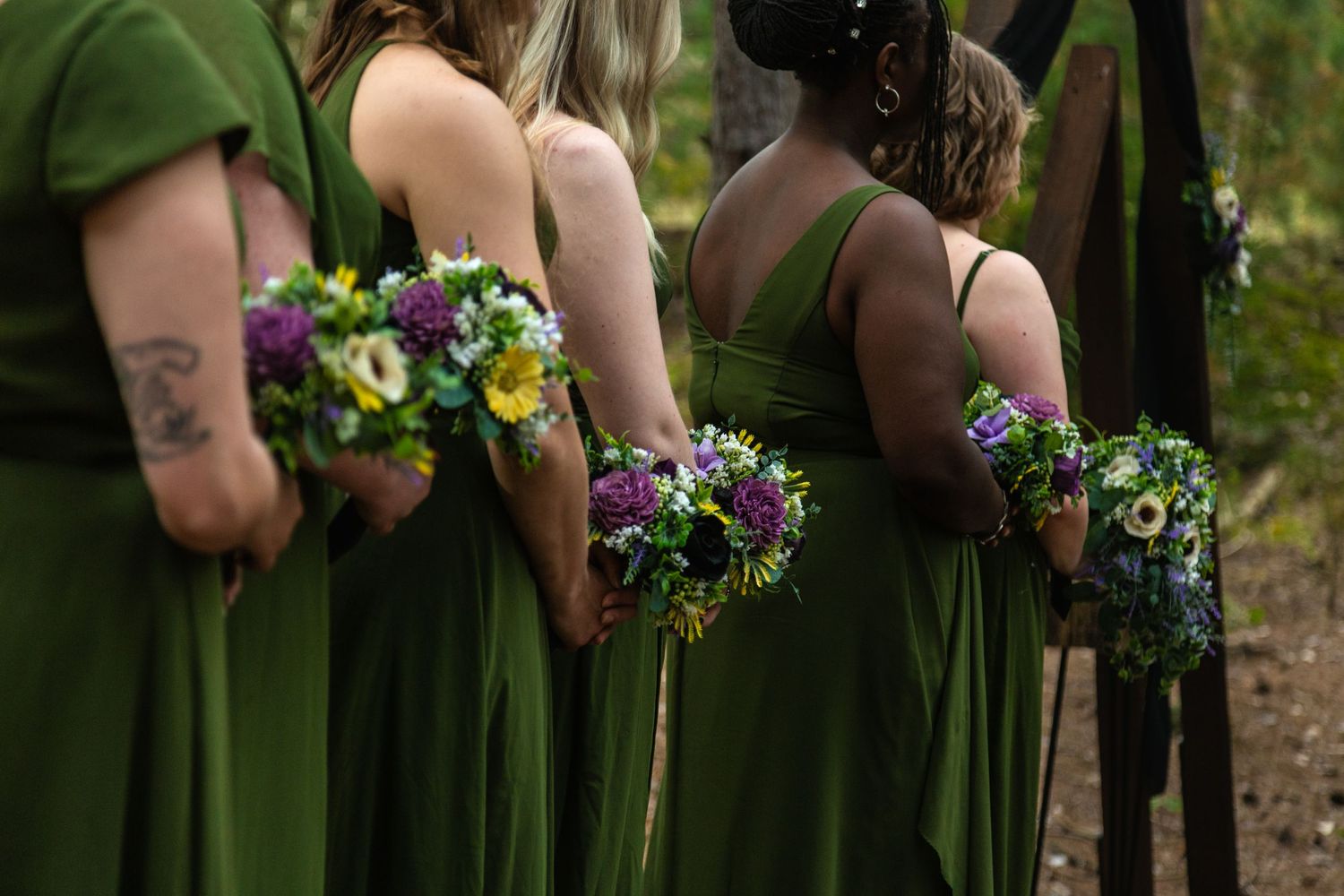 Bridesmaids in emerald green dresses hold colorful floral bouquets while standing in line at an outdoor wedding. Ludington, Michigan.
