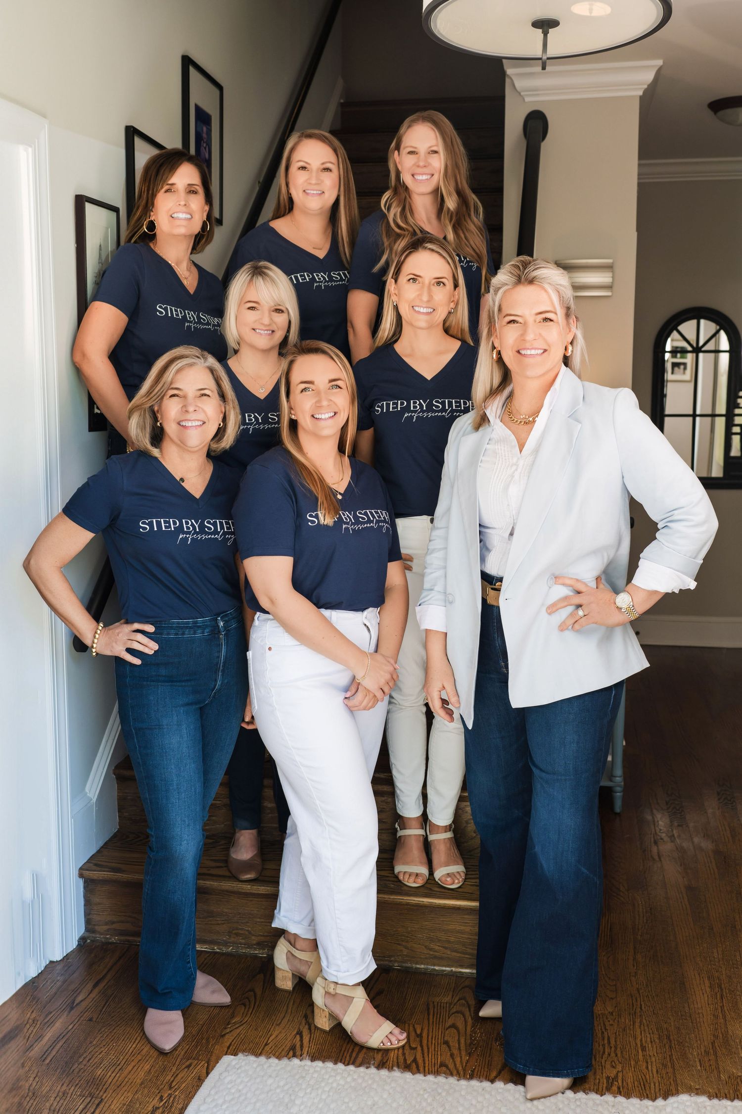 Group of women wearing matching navy blue shirts and jeans pose together on wooden stairs in a home interior. branding