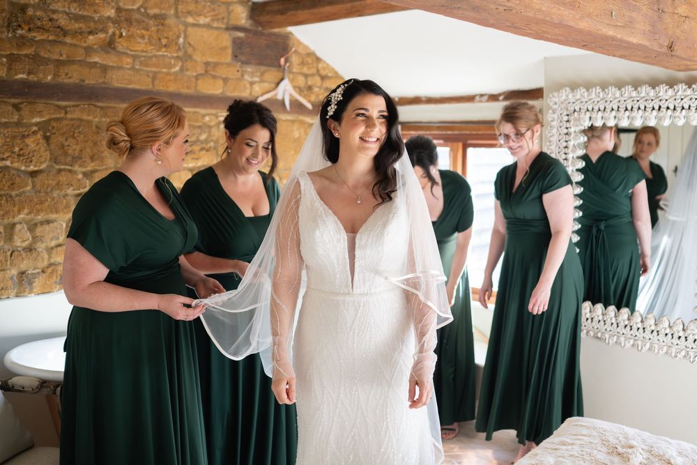 Bridesmaids in emerald green dresses help the bride prepare in a rustic stone-walled room.