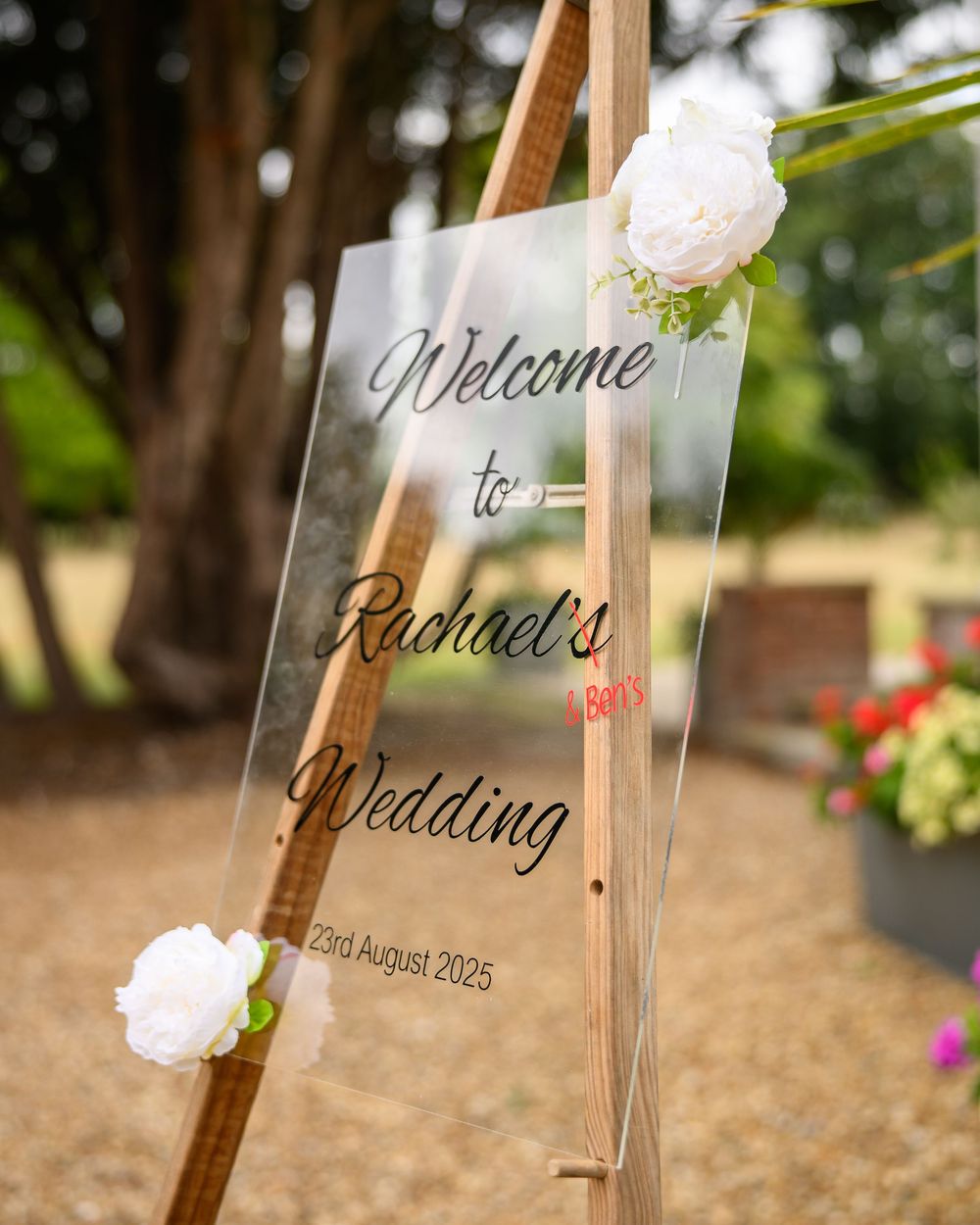 Clear acrylic welcome sign with white flowers on a wooden easel displays elegant wedding details in an outdoor garden setting.