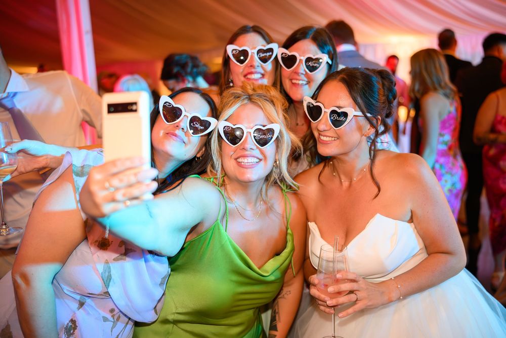 Wedding party guests wearing matching sunglasses pose together for fun photos at an indoor reception.