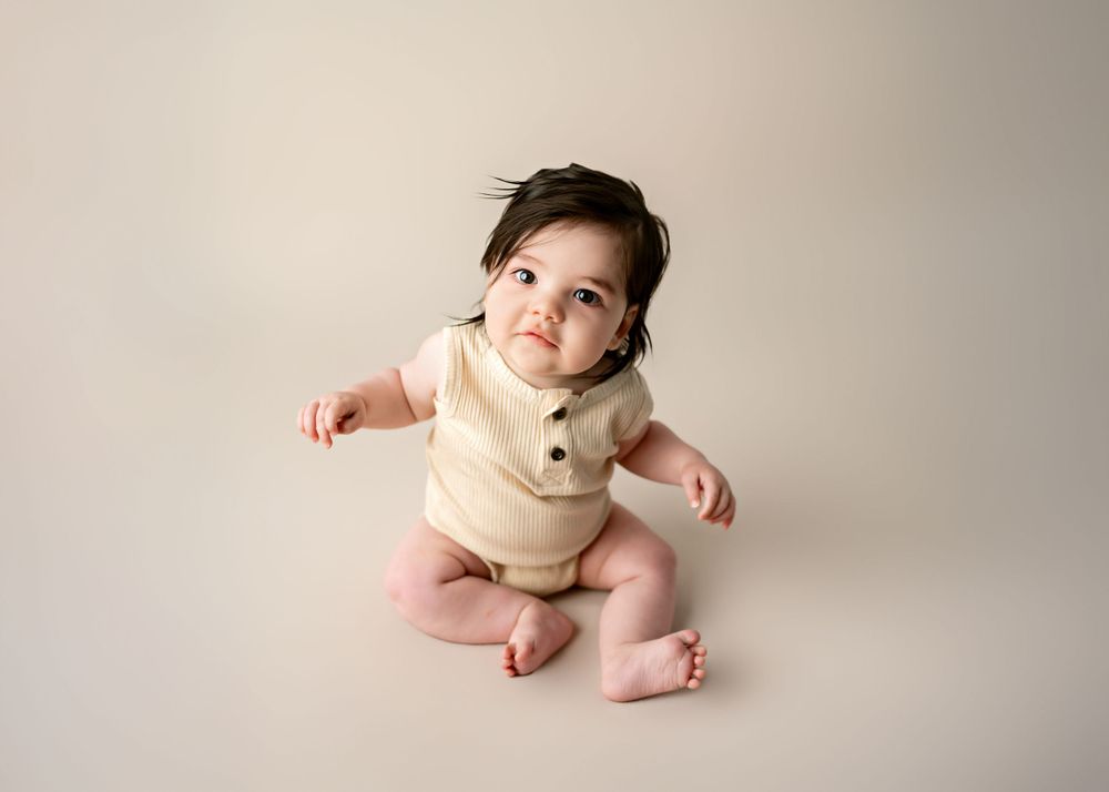 Baby wearing beige knit romper sitting on neutral backdrop during studio photoshoot.