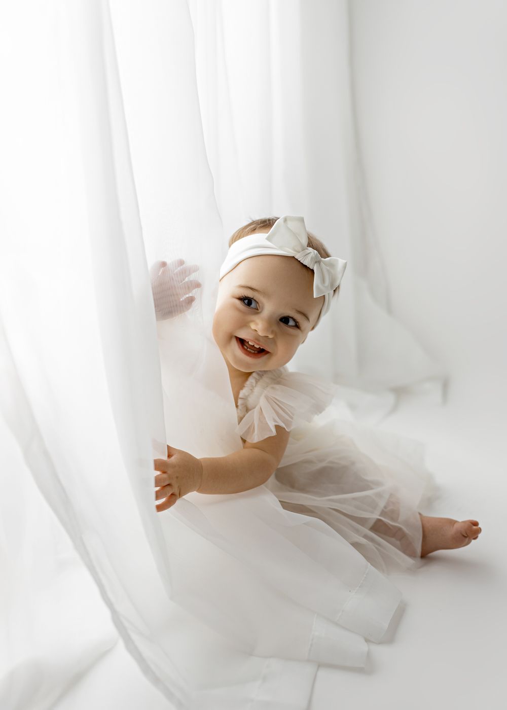 Baby in white tulle dress and headband playing peek-a-boo behind sheer white curtains.