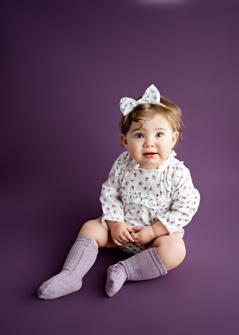 Small child wearing white patterned top with bow and lavender socks sits against purple background during photo session.