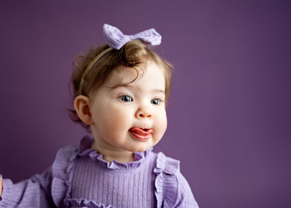 A toddler wearing a purple sweater and bow smiles while looking away against a deep purple backdrop.