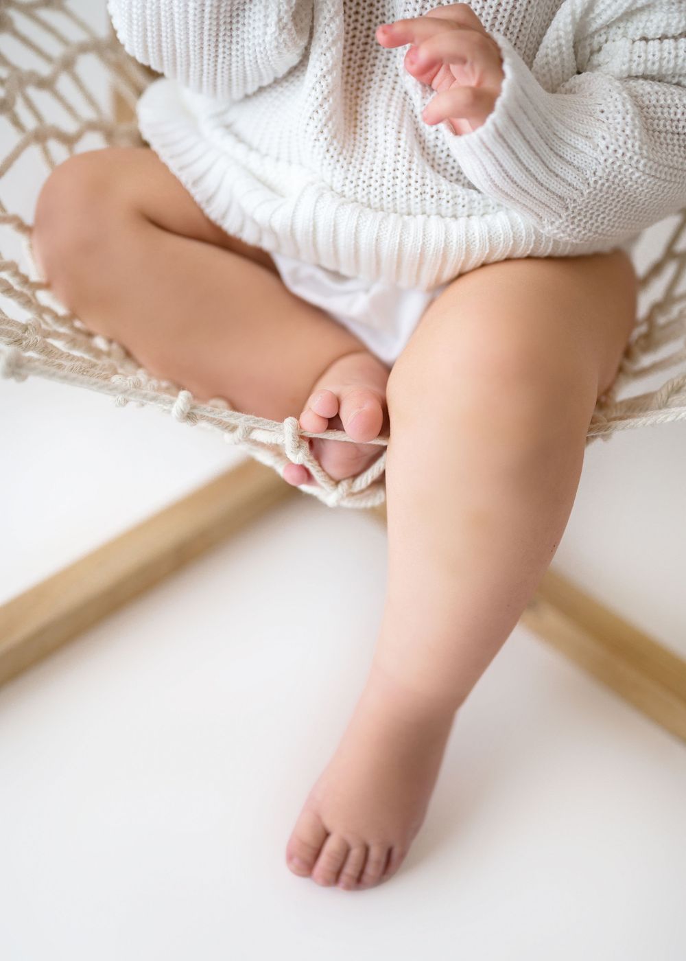 Close-up of baby's legs and feet in white knit sweater sitting on wooden frame.
