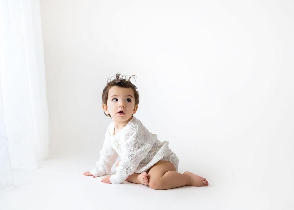 Baby in white outfit crawls on white floor near window.