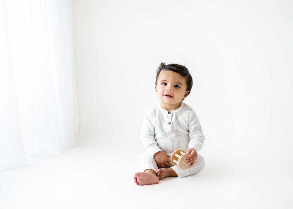 Baby in grey henley shirt sits with snack against white backdrop.