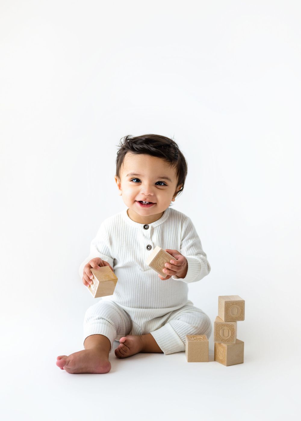 A baby in white ribbed outfit sits with wooden blocks on a white background.