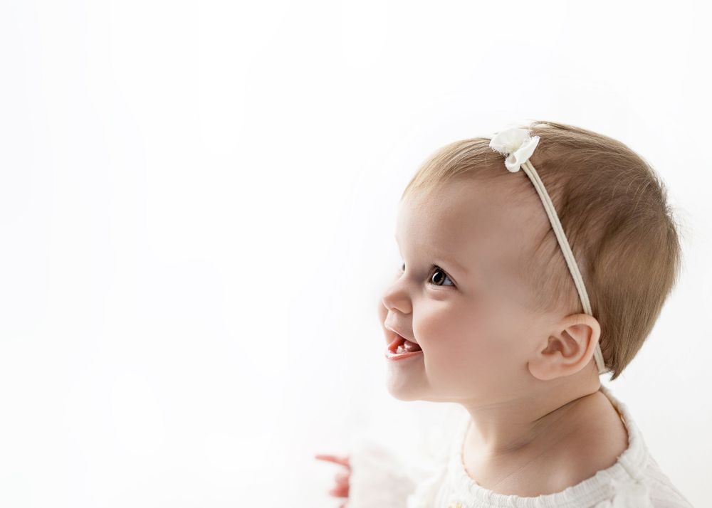 Baby wearing white headband looks up with wonder against white backdrop.
