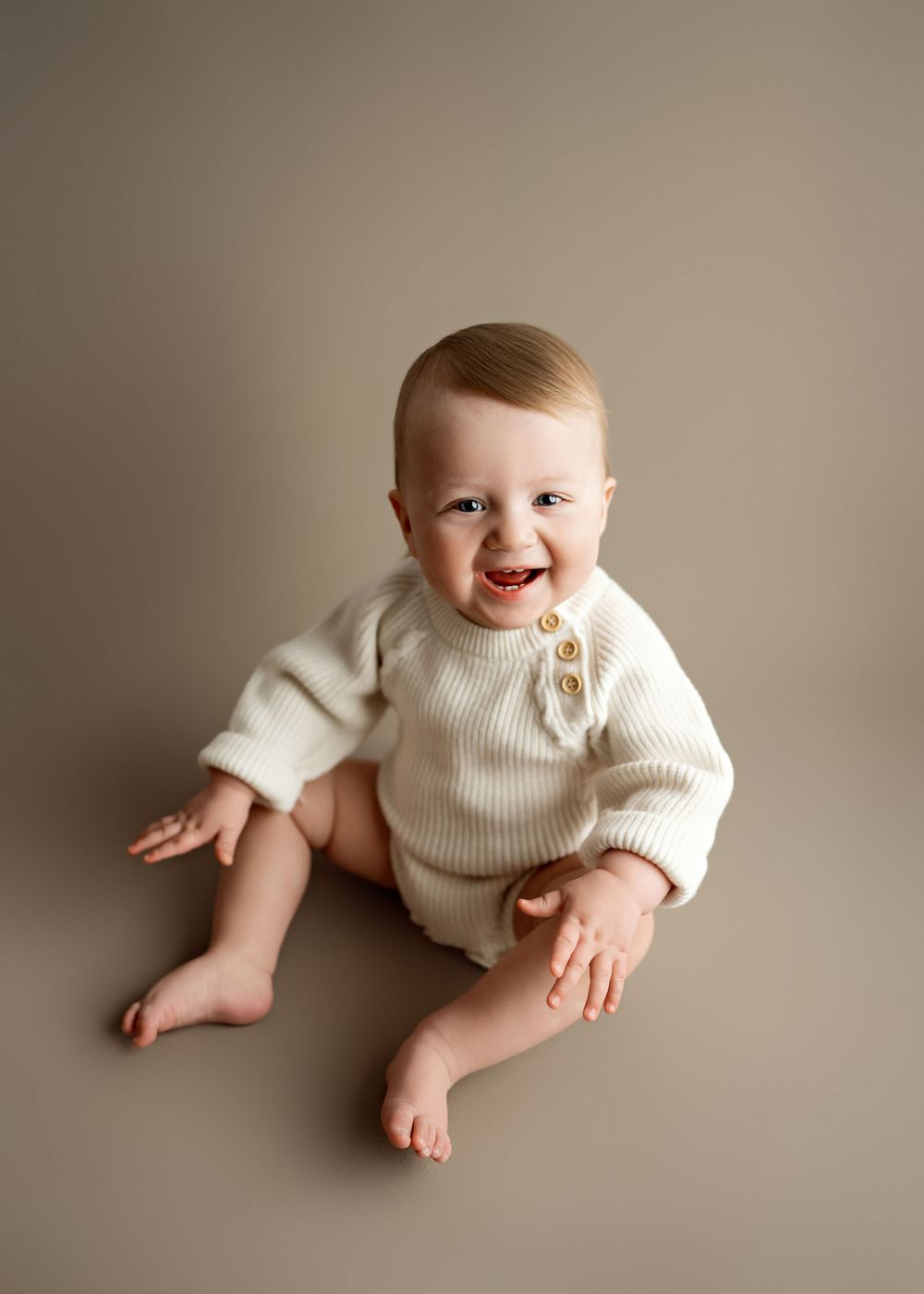 Happy baby wearing cream-colored knit romper sits on neutral beige background showing pure joy and laughter.