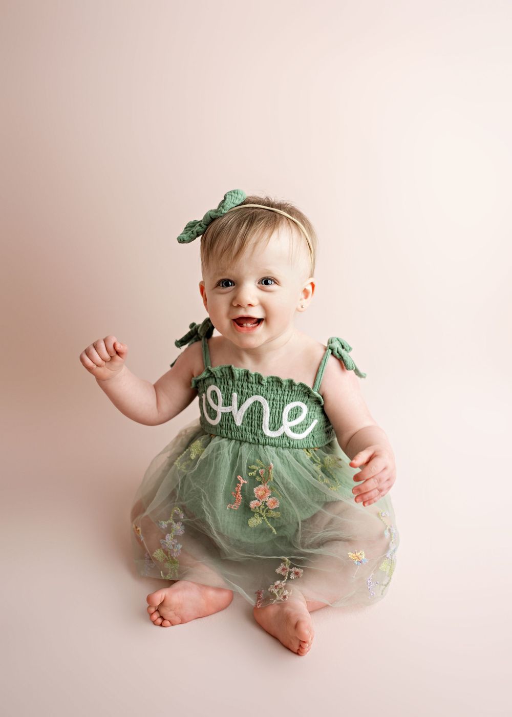 A happy baby in a mint green tulle dress with the word 'one' and floral designs sits against a light pink background.