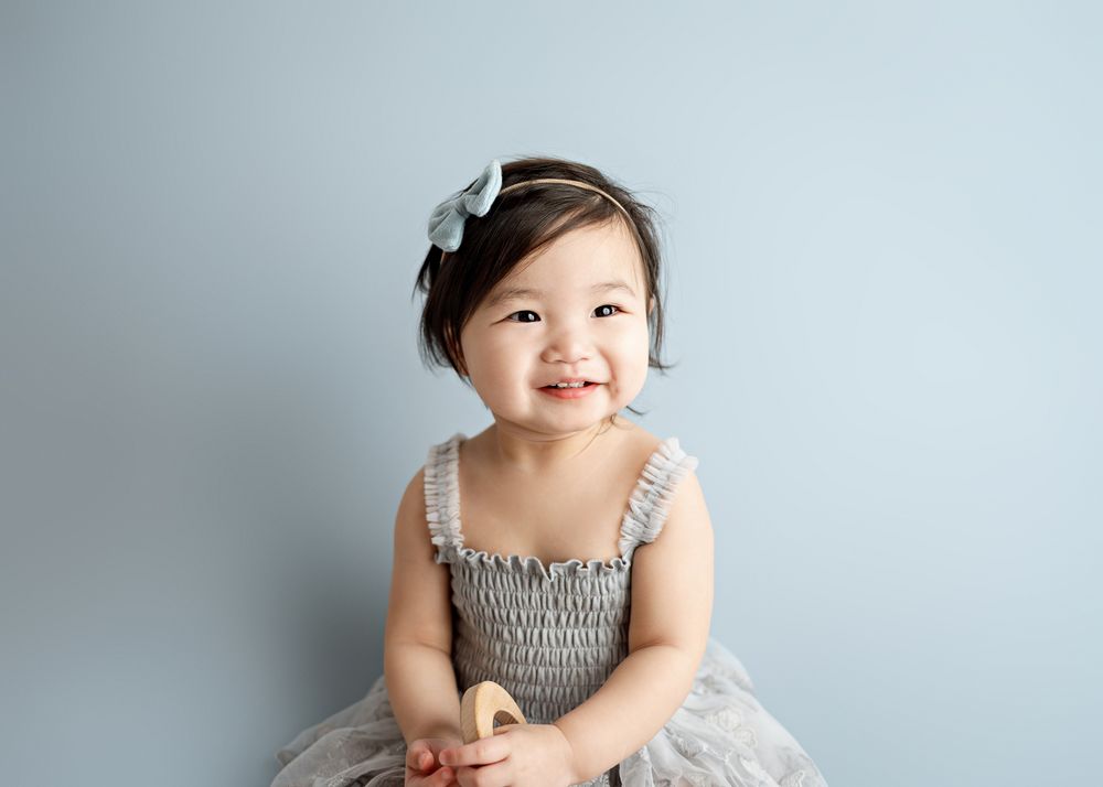 A smiling toddler in a gray tulle dress and hair bow poses against a light blue backdrop.