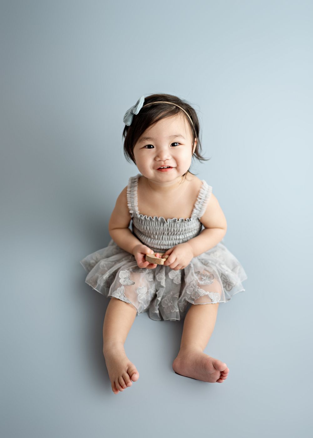 Little one in a gray tulle dress with ruffled top and bow headband smiles sweetly while sitting against light background.
